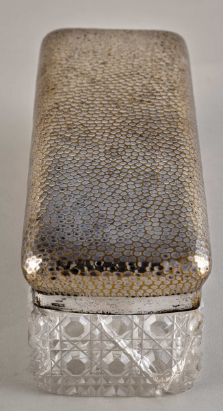 Na pierwszym planie widoczne jest pudełko z posrebrzanym spodem z wzorami oraz pokrywą.