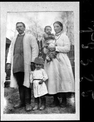 Fotografia czarno-biała przedstawia rodzinę w strojach biłgorajskich. Postacie stoją na tle drewnianego płotu. Od lewej strony: ojciec, kilkuletni syn i matka z niemowlęciem na ręku. 