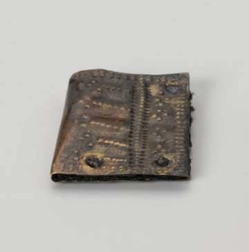 Okucie pochewki - ujęcie z boku; Czworoboczne okucie skórzanej pochewki ze stopu metali, wykonane ze zgiętej w pół blaszki. Mocowane czterema nitami w trzech narożnikach. Zdobione ornamentem punktowym wybijanym po obu stronach okucia. Na spodniej stronie podwójne punktowanie biegnie wzdłuż krawędzi, na stronie wierzchniej oprócz analogicznego zdobienia przy krawędzi pojawia się także bardziej skomplikowany układ kropek na całej powierzchni, złożony z linii prostych i wybijanych punktów układających się w czworoboki. Między warstwami blaszki zachowały się fragmenty skóry i włókien tkaniny.