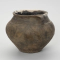 Waza typu IV (Wołągiewicz) - ujęcie z boku; Naczynie ceramiczne (waza) barwy ciemno brunatnej o chropowaconej części dolnej i wygładzanej górnej partii. Waza ma dwa przewężone ucha. Na brzuścu zdobiona chropowaconymi pasami w układzie podwójnego zygzaka. Wysokość wazy wynosi 19,8 cm, średnica brzuśca 22 cm, a średnica wylewu 21 cm.