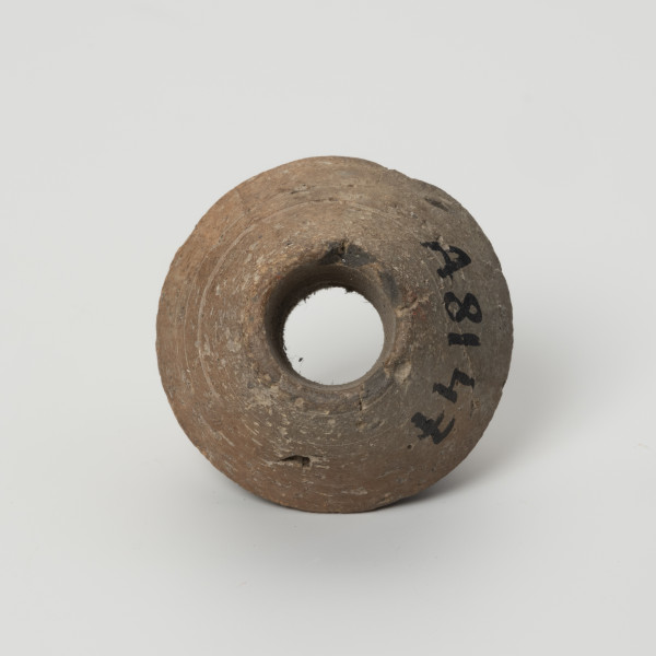 Przęślik dwustożkowy - ujęcie z dołu; Przęślik gliniany dwustożkowy o ściętych, płaskich biegunach, dobrze wypalony, o barwie jasnobrązowej. Na powierzchni widoczne ślady toczenia oraz miejscami ubytki gliny. Wzdłuż osi pionowej przęślika znajduje się otwór o średnicy ok. 0,8 cm. Jeden z biegunów nieznacznie uszkodzony.