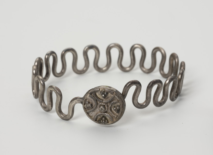 Bransoleta wężykowata - ujęcie z przodu; Bransoleta srebrna wężykowata z okrągłym zapięciem kapsułkowym z lutowanej blachy. Zapięcie zdobione jest filigranem oraz drutem perełkowym. Średnica zamknięcia wynosi 1,4 cm. Kabłąk z kolei tworzy srebrny drut o średnicy 2 mm wygięty w gęsty esowaty zygzak.