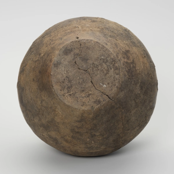 Waza typu IV (Wołągiewicz) - ujęcie z dołu; Naczynie ceramiczne (waza) barwy ciemno brunatnej o chropowaconej części dolnej i wygładzanej górnej partii. Waza ma dwa przewężone ucha. Na brzuścu zdobiona chropowaconymi pasami w układzie podwójnego zygzaka. Wysokość wazy wynosi 19,8 cm, średnica brzuśca 22 cm, a średnica wylewu 21 cm.