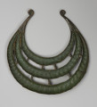 Naszyjnik kołnierzowaty - ujęcie z dołu; Brązowy naszyjnik kołnierzowaty zwany także napierśnikiem składa się z trzech półksiężycowatych obręczy o różnej średnicy, zestawionych w koncentryczny układ. Ich końce schodzą się ze sobą, tworząc wspólne zakończenie w formie niewielkich wolutowych uszek. W wolnych przestrzeniach między kręgami znajdują się krótkie, proste poprzeczne łączniki: 5 między zewnętrzną i środkową obręczą i 4 między środkową i wewnętrzną. Kręgi mają przekrój poprzeczny C-kształtny. Powierzchnie ozdoby dekoruje ukośne żłobkowanie imitujące skręcony sznur. Na największej zewnętrznej obręczy, w pobliżu łączenia z innymi, widoczne są pasma ukośnych nacięć, po siedem z każdej strony. Na obydwu końcach naszyjnika wyryto motywy trójkąta. Barwa ciemnobrązowa ze śladami zielonej patyny w zagłębieniach żłobkowania, na jednym z końców w obszarze ukośnych nacięć złotawa.
