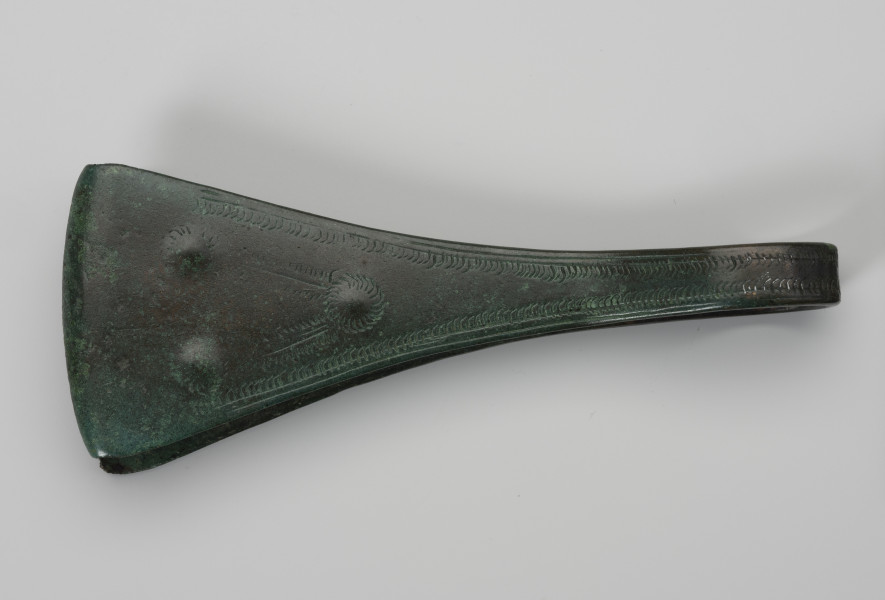 Pinceta - ujęcie z góry; Dość masywna pinceta z brązu o wyraźnie przewężonym kabłąku przechodzącym w szerokie trójkątne ramiona. Brzegi kabłąka są zdobione drobnymi nacięciami i podwójną linią rytą. Natomiast po środku ramion trzy niewielkie ozdobne guzki wyznaczają trójkąt, którego dwa boki tworzy liniowy ornament w postaci motywu tzw. jodełki. Powierzchnia zabytku pokrywa zielona szlachetna patyna.