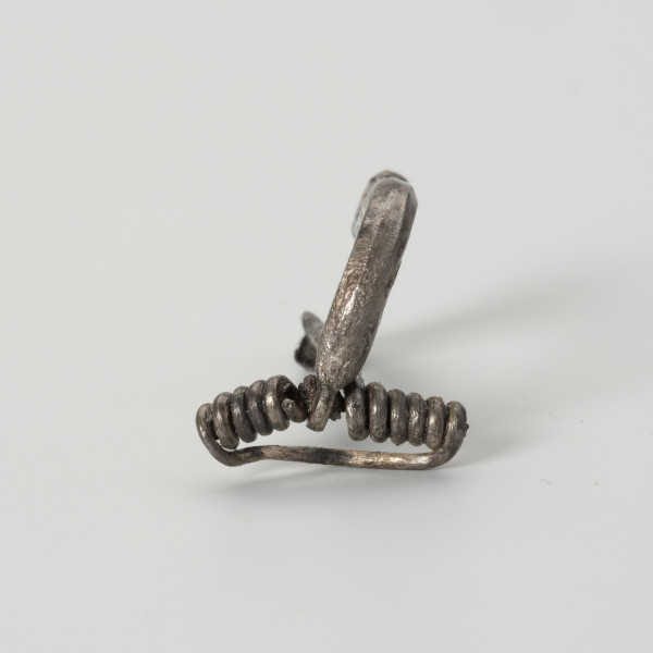 Fibula typu VII - ujęcie z przodu; Fibula srebrna z wysoką pochewką o wysokości 1,2 cm i szerokość sprężynki 1,8 cm. Zapinka posiada zdobienie w postaci niewielkiego guzka na końcu kabłąka. Fibula odpowiadająca VII grupie w typologii Oscara Almgrena