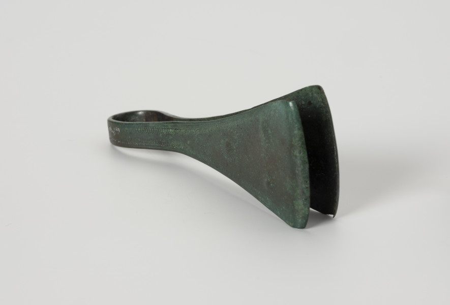 Pinceta - ujęcie z boku; Dość masywna pinceta z brązu o wyraźnie przewężonym kabłąku przechodzącym w szerokie trójkątne ramiona. Brzegi kabłąka są zdobione drobnymi nacięciami i podwójną linią rytą. Natomiast po środku ramion trzy niewielkie ozdobne guzki wyznaczają trójkąt, którego dwa boki tworzy liniowy ornament w postaci motywu tzw. jodełki. Powierzchnia zabytku pokrywa zielona szlachetna patyna.