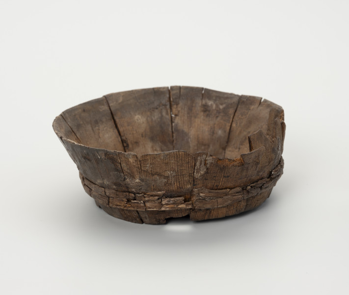 Miska klepkowa - ujęcie z boku; Miska klepkowa zachowana w całości, nieco uszkodzona i zdeformowana, zakonserwowana, zbudowana z wielu elementów: dna, 10 klepek oraz obręczy spajającej klepki złożonej z podwójnego zwoju łyka. Dno koliste, ze ściętą ukośnie krawędzią, umieszczoną w wyciętych specjalnie w dolnej części wewnętrznych stron klepek, otworach, zwanych wątorami. Klepki spojone zostały obręczą z łyka, umieszczoną w zagłębieniach wyciętych w zewnętrznych ich stronach.