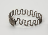 Bransoleta wężykowata - ujęcie ze skosu; Bransoleta srebrna wężykowata z okrągłym zapięciem kapsułkowym z lutowanej blachy. Zapięcie zdobione jest filigranem oraz drutem perełkowym. Średnica zamknięcia wynosi 1,4 cm. Kabłąk z kolei tworzy srebrny drut o średnicy 2 mm wygięty w gęsty esowaty zygzak.