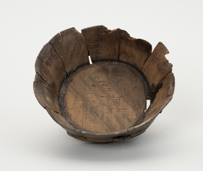 Miska klepkowa - ujęcie z góry; Miska klepkowa zachowana w całości, nieco uszkodzona i zdeformowana, zakonserwowana, zbudowana z wielu elementów: dna, 10 klepek oraz obręczy spajającej klepki złożonej z podwójnego zwoju łyka. Dno koliste, ze ściętą ukośnie krawędzią, umieszczoną w wyciętych specjalnie w dolnej części wewnętrznych stron klepek, otworach, zwanych wątorami. Klepki spojone zostały obręczą z łyka, umieszczoną w zagłębieniach wyciętych w zewnętrznych ich stronach. Spojenia klepek z dnem zostały od strony wewnętrznej uszczelnione czarną substancją, zapewne pochodną dziegciu.