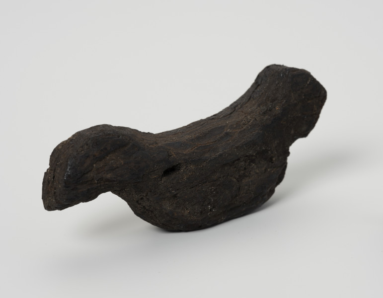 Ptaszek z kory - ujęcie z przodu; Figurka ptaszka wystrugana z kory o schematycznej sylwetce. Wyodrębniona została jedynie głowa i ogon bez pozostałych typowych cech ptasich. Powierzchnia wykończona starannie, bez zadziorów.