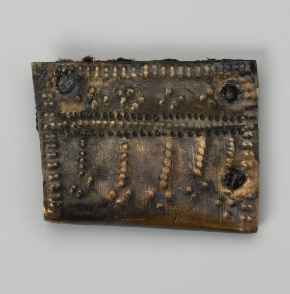 Okucie pochewki - ujęcie z góry; Czworoboczne okucie skórzanej pochewki ze stopu metali, wykonane ze zgiętej w pół blaszki. Mocowane czterema nitami w trzech narożnikach. Zdobione ornamentem punktowym wybijanym po obu stronach okucia. Na spodniej stronie podwójne punktowanie biegnie wzdłuż krawędzi, na stronie wierzchniej oprócz analogicznego zdobienia przy krawędzi pojawia się także bardziej skomplikowany układ kropek na całej powierzchni, złożony z linii prostych i wybijanych punktów układających się w czworoboki. Między warstwami blaszki zachowały się fragmenty skóry i włókien tkaniny.