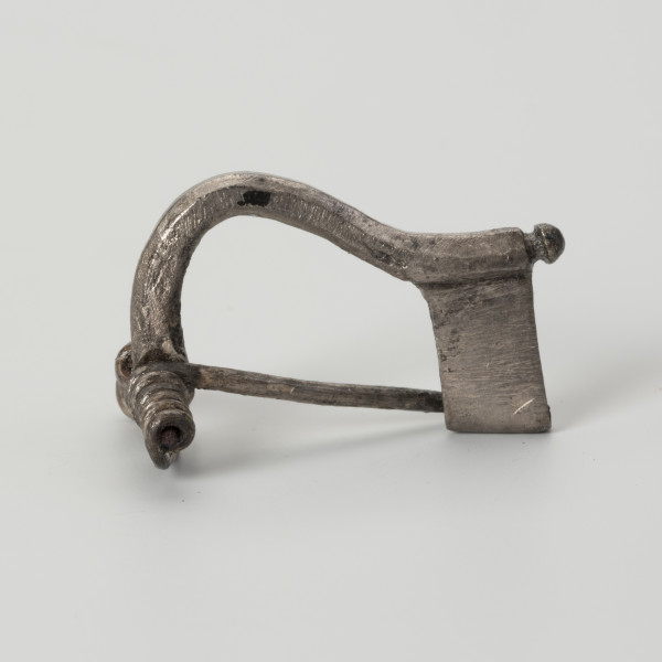 Fibula typu VII - ujęcie z drugiego boku; Fibula srebrna z wysoką pochewką o wysokości 1,2 cm i szerokość sprężynki 1,8 cm. Zapinka posiada zdobienie w postaci niewielkiego guzka na końcu kabłąka. Fibula odpowiadająca VII grupie w typologii Oscara Almgrena