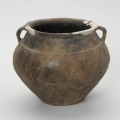 Waza typu IV (Wołągiewicz) - ujęcie z tyłu; Naczynie ceramiczne (waza) barwy ciemno brunatnej o chropowaconej części dolnej i wygładzanej górnej partii. Waza ma dwa przewężone ucha. Na brzuścu zdobiona chropowaconymi pasami w układzie podwójnego zygzaka. Wysokość wazy wynosi 19,8 cm, średnica brzuśca 22 cm, a średnica wylewu 21 cm.