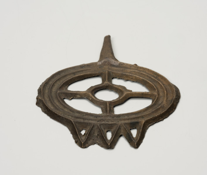 Szpila typu lüneburskiego z główką w kształcie koła szprychowego - ujęcie z tyłu; Jest to szpila z brązu o okazałej kolistej główce w formie tzw. koła szprychowego - tzn. w zewnętrzy, szeroki profilowany okrąg (profilowanie stanowi imitacje trzech współśrodkowych okręgów) wpisany jest mniejszy okrąg, połączony czterema 