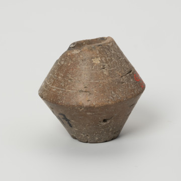 Przęślik dwustożkowy - ujęcie z boku; Przęślik gliniany dwustożkowy o ściętych, płaskich biegunach, dobrze wypalony, o barwie jasnobrązowej. Na powierzchni widoczne ślady toczenia oraz miejscami ubytki gliny. Wzdłuż osi pionowej przęślika znajduje się otwór o średnicy ok. 0,8 cm. Jeden z biegunów nieznacznie uszkodzony.