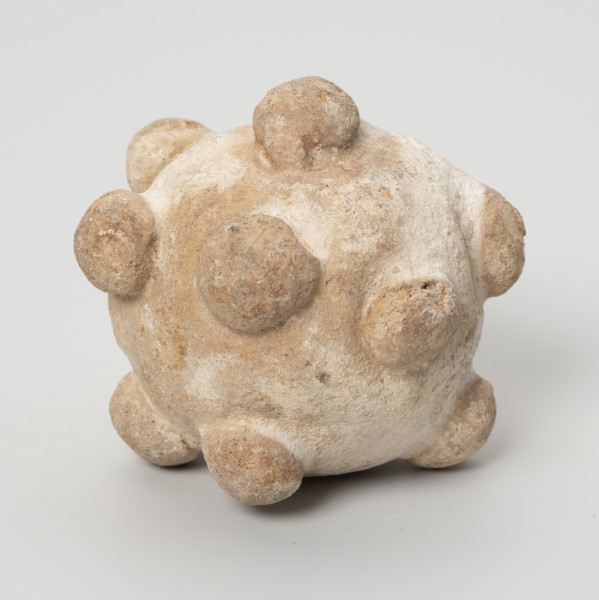 Grzechotka gliniana - ujęcie z boku; Biała okrągła grzechotka gliniana z półkolistymi guzkami i pojedyńczym kamyczkiem w środku.