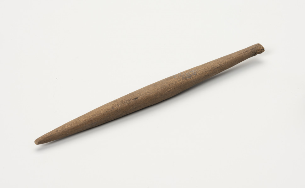 Wrzeciono - ujęcie z boku; Podłużne, smukłe wrzeciono wykonane z drewna. Jest okrągłe w przekroju, zwężające się ku końcom.