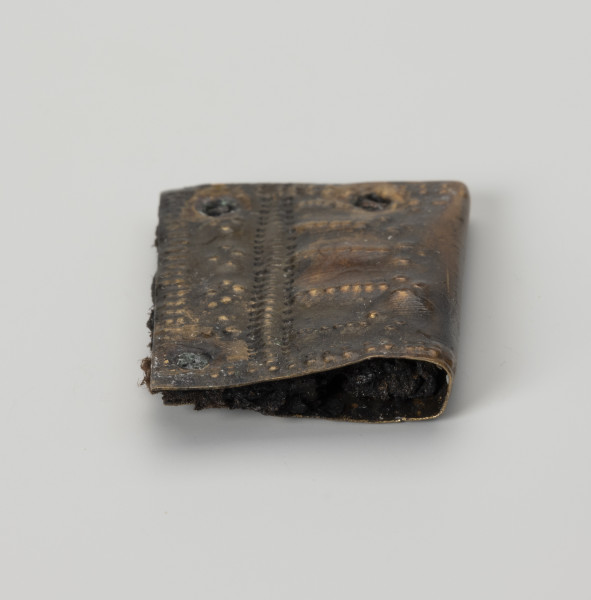 Okucie pochewki - ujęcie z boku; Czworoboczne okucie skórzanej pochewki ze stopu metali, wykonane ze zgiętej w pół blaszki. Mocowane czterema nitami w trzech narożnikach. Zdobione ornamentem punktowym wybijanym po obu stronach okucia. Na spodniej stronie podwójne punktowanie biegnie wzdłuż krawędzi, na stronie wierzchniej oprócz analogicznego zdobienia przy krawędzi pojawia się także bardziej skomplikowany układ kropek na całej powierzchni, złożony z linii prostych i wybijanych punktów układających się w czworoboki. Między warstwami blaszki zachowały się fragmenty skóry i włókien tkaniny.