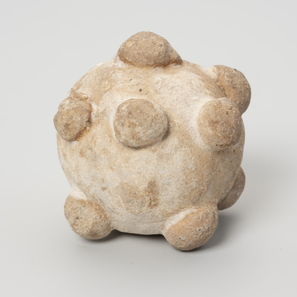 Grzechotka gliniana - ujęcie z przodu; Biała okrągła grzechotka gliniana z półkolistymi guzkami i pojedyńczym kamyczkiem w środku.