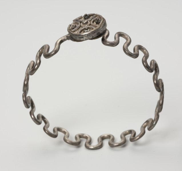 Bransoleta wężykowata - ujęcie z dołu; Bransoleta srebrna wężykowata z okrągłym zapięciem kapsułkowym z lutowanej blachy. Zapięcie zdobione jest filigranem oraz drutem perełkowym. Średnica zamknięcia wynosi 1,4 cm. Kabłąk z kolei tworzy srebrny drut o średnicy 2 mm wygięty w gęsty esowaty zygzak.