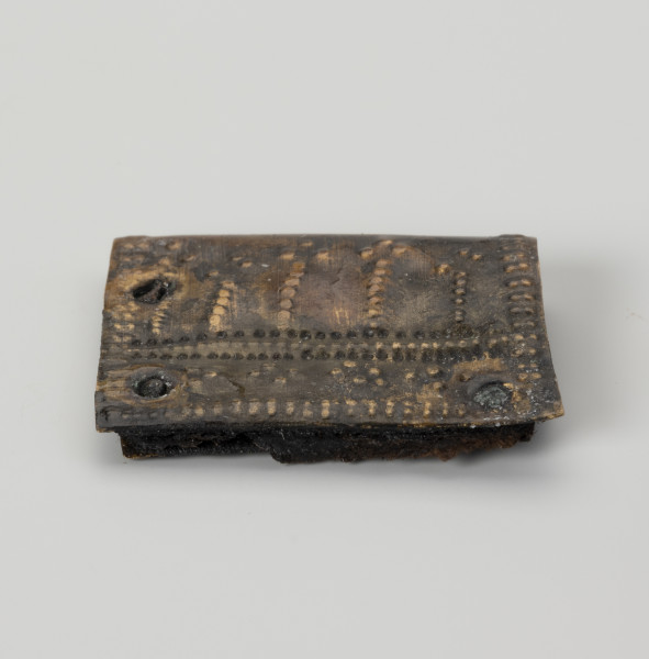 Okucie pochewki - ujęcie z góry; Czworoboczne okucie skórzanej pochewki ze stopu metali, wykonane ze zgiętej w pół blaszki. Mocowane czterema nitami w trzech narożnikach. Zdobione ornamentem punktowym wybijanym po obu stronach okucia. Na spodniej stronie podwójne punktowanie biegnie wzdłuż krawędzi, na stronie wierzchniej oprócz analogicznego zdobienia przy krawędzi pojawia się także bardziej skomplikowany układ kropek na całej powierzchni, złożony z linii prostych i wybijanych punktów układających się w czworoboki. Między warstwami blaszki zachowały się fragmenty skóry i włókien tkaniny.