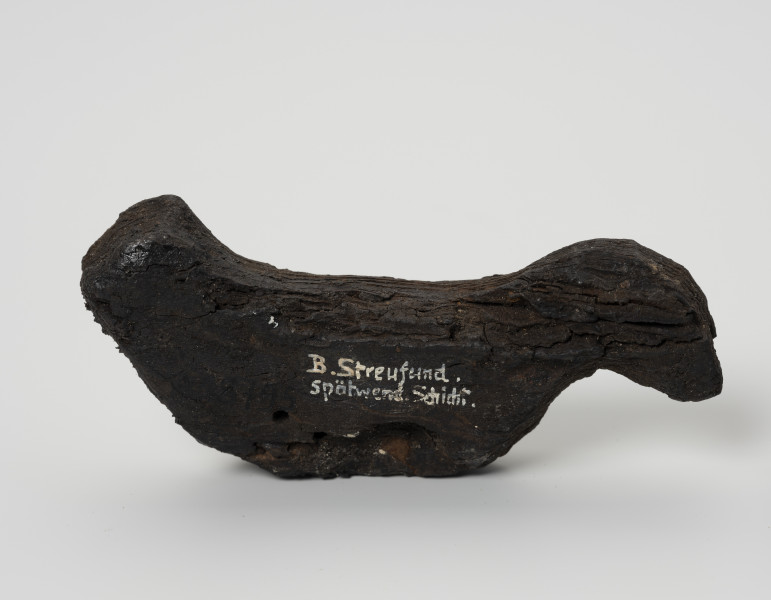 Ptaszek z kory - ujęcie z tyłu; Figurka ptaszka wystrugana z kory o schematycznej sylwetce. Wyodrębniona została jedynie głowa i ogon bez pozostałych typowych cech ptasich. Powierzchnia wykończona starannie, bez zadziorów.