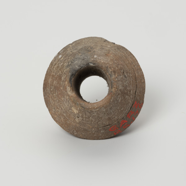 Przęślik dwustożkowy - ujęcie z góry; Przęślik gliniany dwustożkowy o ściętych, płaskich biegunach, dobrze wypalony, o barwie jasnobrązowej. Na powierzchni widoczne ślady toczenia oraz miejscami ubytki gliny. Wzdłuż osi pionowej przęślika znajduje się otwór o średnicy ok. 0,8 cm. Jeden z biegunów nieznacznie uszkodzony.