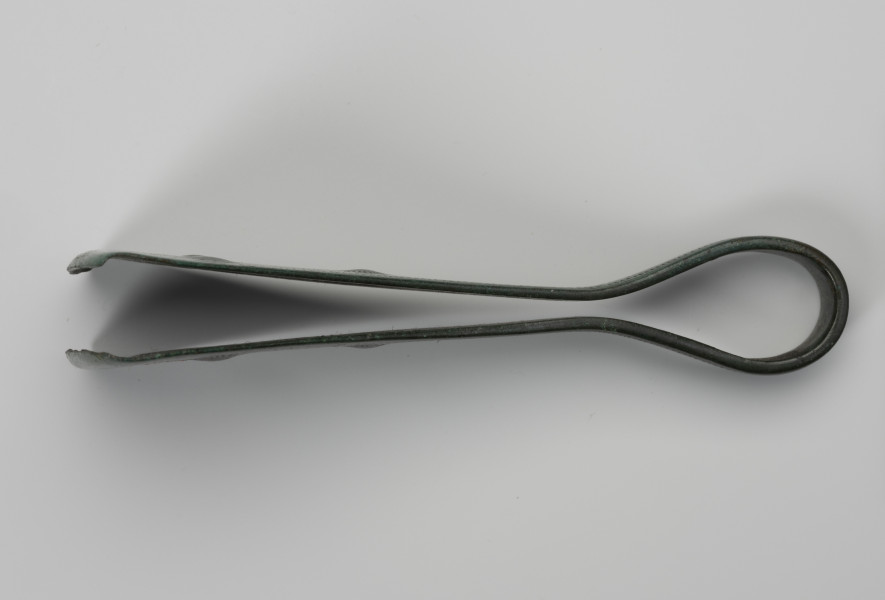 Pinceta - ujęcie z boku; Dość masywna pinceta z brązu o wyraźnie przewężonym kabłąku przechodzącym w szerokie trójkątne ramiona. Brzegi kabłąka są zdobione drobnymi nacięciami i podwójną linią rytą. Natomiast po środku ramion trzy niewielkie ozdobne guzki wyznaczają trójkąt, którego dwa boki tworzy liniowy ornament w postaci motywu tzw. jodełki. Powierzchnia zabytku pokrywa zielona szlachetna patyna.