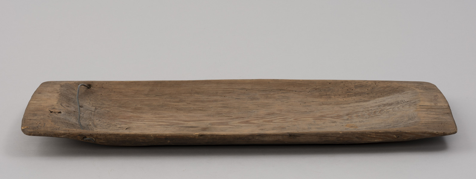 tacka do sznurów haczykowych - ujęcie z boku; Płytka, drewniana niecka, wykonana techniką stolarską z jednego kawałka drewna. Naroża lekko zaokrąglone. Na jednym z końców niecki, przy krawędziach dłuższych boków, po jednym otworze, przez który przechodzi drut, służący do mocowania haczyków.