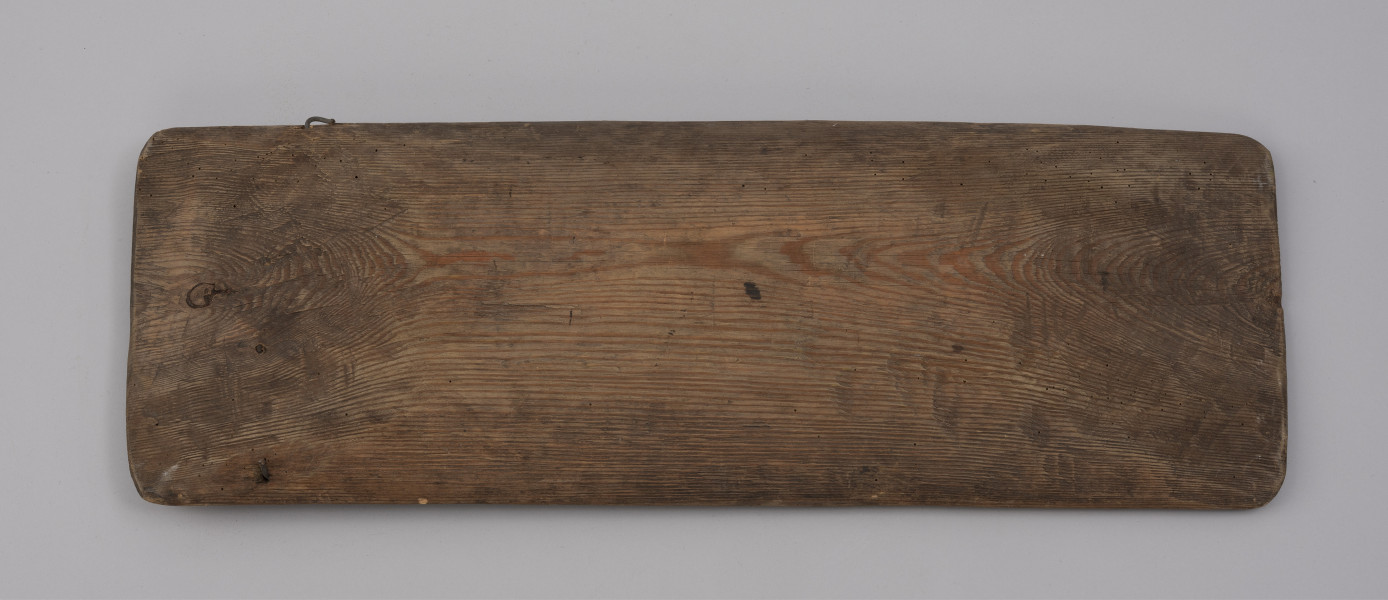 tacka do sznurów haczykowych - ujęcie z dołu; Drewniana niecka, wykonana techniką stolarską z jednego kawałka drewna. Naroża lekko zaokrąglone. Na jednym z końców niecki, przy krawędziach dłuższych boków, po jednym otworze, przez który przechodzi drut, służący do mocowania haczyków.