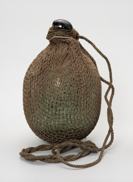Pływak - ujęcie z przodu; Wykonany z ciemnozielonego szkła pływak o kształcie przypominającym kulę. W górnej części pływak przechodzi w krótką, zakończoną spłaszczoną gałką szyjkę. Pływak w całości dwukrotnie owinięty siatką bawełnianą o małych, kwadratowych oczkach. Końce siatki ściśle przylegają do szyjki i są mocowane do niej za pomocą kilkukrotnie owiniętego wokół szyjki sznurka. Drugi, grubszy sznurek, służący jako mocowanie do sieci również owinięty jest wokół szyjki.