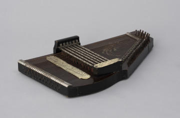 Cytra klawiszowa (autoharp) - ujęcie z prawej; Cytra klawiszowa (autoharp) z płaskim pudłem rezonansowym wykonanym z drewna polichromowanego o kształcie przypominającym trapez z jednym ściętym rogiem. Wzdłuż najkrótszego boku oraz boku odchodzącego od niego pod kontem rozwartym, szereg metalowych kołków, do których przymocowanych jest 25, diatonicznie strojonych strun, ułożonych od najgrubszej – górnej – do najcieńszej z dolnej strony instrumentu; ostatnie dwie są zerwane. Poniżej kołków znajdują się dwie, zestawione pod kątem rozwartym, wąskie, metalowe blaszki z niewielkimi bolcami, stanowiące wsparcie i gwarantujące właściwy rozstaw strun. Podobna blaszka zamontowana jest wzdłuż dolnej krawędzi instrumentu. Struny przechodzą przez znajdujące się w niej rowki, zaginają pod kątem 90 stopni i mocowane są w znajdujących się w bocznej ściance instrumentu bolcach, ułożonych naprzemiennie: wyżej – niżej. Bezpośrednio nad strunami, prostopadle względem nich, znajduje się sześć metalowych prętów zamontowanych pomiędzy dwoma drewnianymi mostkami. Osadzone na sprężynach pręty wprawiane są w ruch przez grającego za pomocą znajdujących się w ich górnej części przycisków. Powoduje to dociśnięcie do struny mocowanych na prętach klawiszy z filcowymi podkładkami. Na jednym z mostków, na przykręconej za pomocą dwóch śrub plakietce z papieru welinowego, znajdują się oznaczenia akordów. Druga plakietka, z zapisem nut w formie literowej i na pięciolinii oraz ciągiem cyfr rzymskich i arabskich, umieszczona jest bezpośrednio na pudle rezonansowym, poniżej mechanizmu klawiszowego. Cytra ozdobiona jest w górnej części kalkomanią.