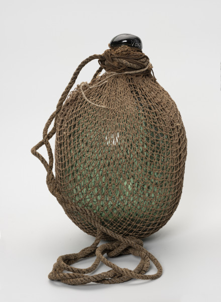 Pływak - ujęcie z przodu; Wykonany z ciemnozielonego szkła pływak o kształcie przypominającym kulę. W górnej części pływak przechodzi w krótką, zakończoną spłaszczoną gałką szyjkę. Pływak w całości dwukrotnie owinięty siatką bawełnianą o małych, kwadratowych oczkach. Końce siatki ściśle przylegają do szyjki i są mocowane do niej za pomocą kilkukrotnie owiniętego wokół szyjki sznurka. Drugi, grubszy sznurek, służący jako mocowanie do sieci również owinięty jest wokół szyjki.
