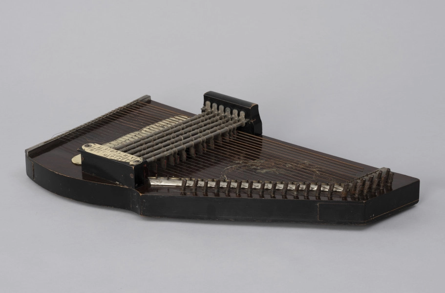 Cytra klawiszowa (autoharp) - ujęcie z boku; Cytra klawiszowa (autoharp) z płaskim pudłem rezonansowym wykonanym z drewna polichromowanego o kształcie przypominającym trapez z jednym ściętym rogiem. Wzdłuż najkrótszego boku oraz boku odchodzącego od niego pod kontem rozwartym, szereg metalowych kołków, do których przymocowanych jest 25, diatonicznie strojonych strun, ułożonych od najgrubszej – z prawej – do najcieńszej z lewej strony instrumentu; ostatnie dwie są zerwane. Przed kołkami znajdują się dwie, zestawione pod kątem rozwartym, wąskie, metalowe blaszki z niewielkimi bolcami, stanowiące wsparcie i gwarantujące właściwy rozstaw strun. Podobna blaszka zamontowana jest wzdłuż dolnej krawędzi instrumentu. Struny przechodzą przez znajdujące się w niej rowki. Na środku instrumentu bezpośrednio nad strunami, prostopadle względem nich, znajduje się sześć metalowych prętów zamontowanych pomiędzy dwoma drewnianymi mostkami. Osadzone na sprężynach pręty wprawiane są w ruch przez grającego za pomocą znajdujących się w ich górnej części przycisków. Powoduje to dociśnięcie do struny mocowanych na prętach klawiszy z filcowymi podkładkami. Na jednym z mostków, na przykręconej za pomocą dwóch śrub plakietce z papieru welinowego, znajdują się oznaczenia akordów.