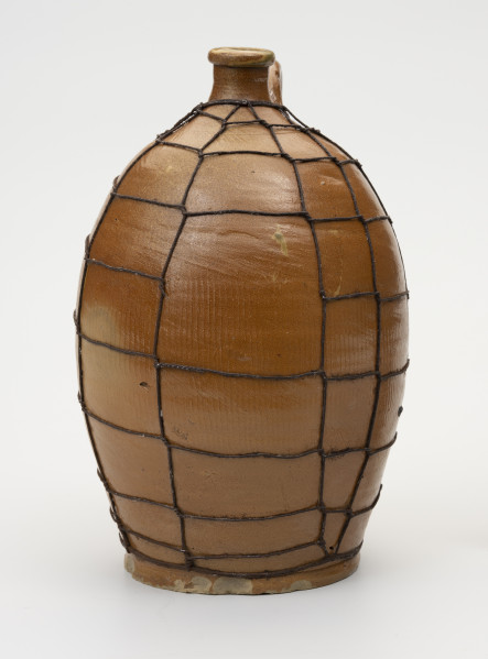 Butla z odrutowaniem - ujęcie z boku; Kamionkowa butla o owalnym kształcie i wyraźnie wyodrębnionym dnie. Brzusiec w dolnej części wyraźnie węższy, o największym rozchyleniu powyżej połowy wysokości, w górnej partii przechodzi bezpośrednio w szyjkę. Szyjka krótka, wąska, wyraźnie wyodrębniona z pogrubionym wylewem. Glazura szyjki na zewnątrz brązowa. Butla odrutowana cienkim drutem. Stopa butli z widocznymi ubytkami.