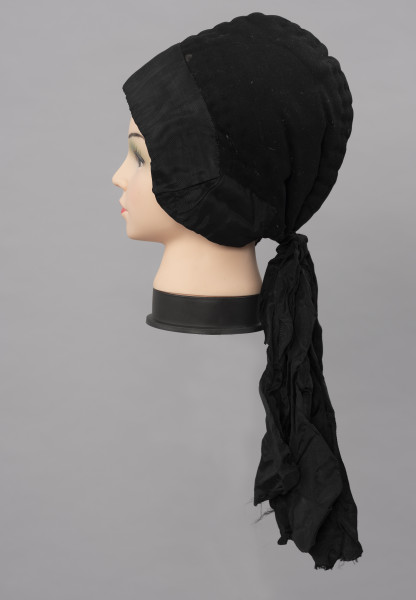 Czepek damski - ujęcie z boku; Czepek damski, codzienny. Wykonany z tkanin produkcji fabrycznej (wełna, atłas) i białego, lnianego płótna wykonanego na krosnach tkackich splotem płóciennym prostym. Główka czepca  z tkaniny wełnianej, czarnej, wokół główki naszyta czarna atłasowa tkanina. Z tyłu dekoracyjna kokarda z atłasowej wstążki. Czepek zszyty z dwóch kawałków, szew wzdłuż pionowej linii głowy, ściągany nad karkiem nicią. Główka wyraźnie zaokrąglona, zachodząca wyraźnie na uszy. Od środka czepek podbity lnianym, białym płótnem i wypełniony grubym usztywniającym materiałem. Przez wszystkie warstwy tkanin przechodzą w wielu miejscach przeszycia białymi i czarnymi nićmi tworzącymi ich złączenie  jako przepikowanie tkaniny.  

Literatura: 