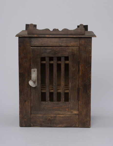 Szafka do przechowywania jajek - ujęcie z przodu; Szafka do przechowywania jajek o konstrukcji ramowej w kształcie prostopadłościanu. Płycina jednoskrzydłowych drzwi wypełnione trzema pionowymi szczebelkami. Drzwiczki zamykane na zapadkę uruchamianą klamką. Wewnątrz widoczne półki z wyciętymi otworami do wkładania jaj. Na desce zamykającej szafkę od góry przybita ozdobnie fazowana listwa.