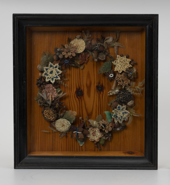 Wieniec pamiątkowy w ramie ze szkłem - ujęcie z przodu; Ozdobny wieniec w drewnianej ramie za szkłem. Rama gruba, profilowana, politurowana na czarno. Wewnątrz ramy, na lakierowanej desce, umieszczony centralnie wieniec wypełnia znaczną część powierzchni. Jego podstawę stanowi metalowy drut, do którego mocowane są elementy dekoracyjne: kwiaty i prawdopodobnie gwiazdki. Wykonane są z ziaren ryżu i gorczycy, z których część jest barwiona na niebiesko lub różowo. Uzupełnienie kompozycji stanowią listki wykonane z zielonej tkaniny i piórek oraz: gwiazdki anyżu, laski cynamonu, migdały, nasiona gałki muszkatołowej, ziarna pieprzu i koraliki.