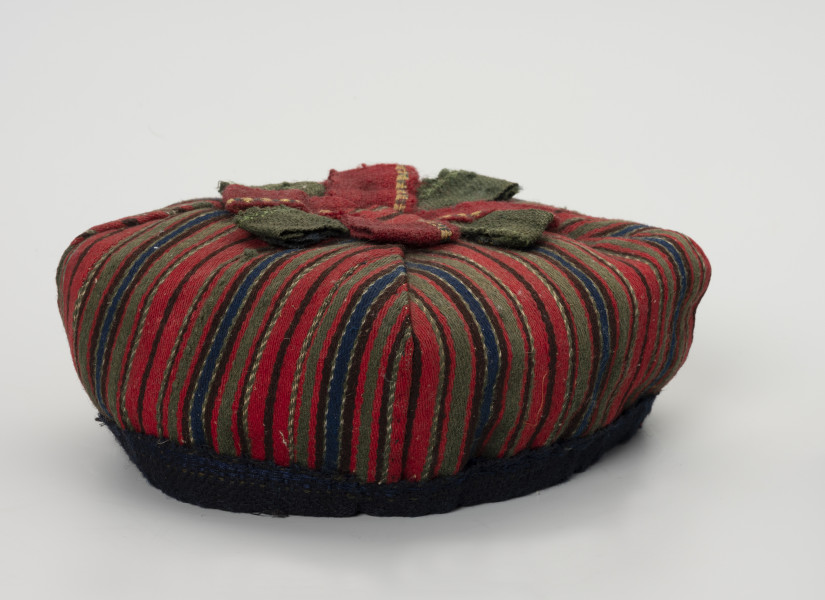 Czapka męska - ujęcie z boku; Czapka męska. Wykonana z tkaniny wełnianej, samodziałowej tzn. na krosnach tkackich splotem skośnym, wzór układa się w paski: czerwony, zielony, granatowy, brązowy, przetykany białym). Czapka ma fason biretu (zbliżonego do nakryć głowy duchownych m.in.  luterańskich). Uszyta jest z  czterech kawałków tkaniny usztywnionych od spodu i zszytych w ten sposób, że kształt zbliżony jest do kwadratu. Na czubku czerwono-zielona pasmanteryjna kokarda płasko ułożona w formę gwiazdy. Otok o zbliżonym do koła obwodzie z granatowej, wełnianej tkaniny.