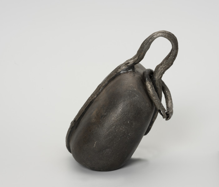 Ciężarek do sieci - ujęcie z boku; Ciężarek do obciążania sieci wykonany z szarego kamienia o gładkiej powierzchni. Kamień otoczony żelaznym, kutym pierścieniem, którego końce nie łączą się ze sobą (wtórne uszkodzenie metalu). W części górnej pierścień wystaje poza obręb kamienia tworząc pętlę, w której zamocowane jest, również wykonane z kutego żelaza, trójkątne ogniwo z zaokrąglonymi kątami i bokami.