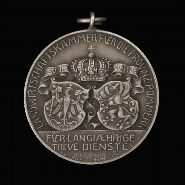 Medal nagrodowy Izby Rolniczej Prowincji Pomorze - awers; Okrągły medal z kompozycją herbową i realistyczną, z uszkiem i koluszkiem. Na awersie herby Królestwa Prus i Prowincji Pomorze, nad nimi korona królewska i szarfa z napisem: SVVM – CVIQVE (każdemu, co mu się należy). Pod tarczą Pomorza monogram MK. W otoku napis: LANDWIRTSCHAFTSKAMMER·FVER·DIE·PROVINZ·POMMERN. W odcinku napis: FVER·LANGIAEHRIGE | TREVE·DIENSTE (za długoletnią wierną służbę). Poniżej  w półkolu sygnatura: L.CHR.LAUER NÜRNBERG.