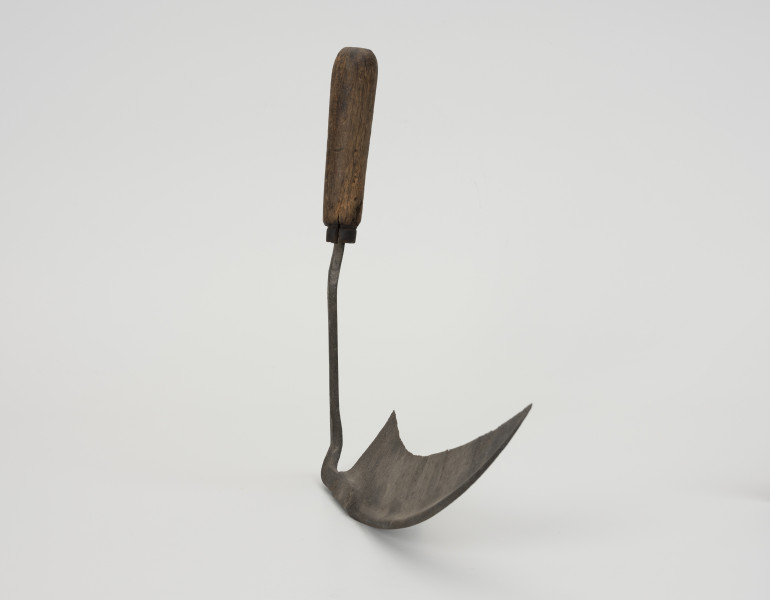 Półkosek - ujęcie z boku; Półkosek składający się z żelaznego ostrza i drewnianego uchwytu  kształtem podobny jest do małej kosy. Żelazne, wydłużone ostrze zwężające się ku końcowi tworzy szpiczaste zakończenie, które osadzone jest na żelaznym kosisku przymocowanym za pomocą dwóch nitów do drewnianej, toczonej rękojeści, zakończonej bolcem.