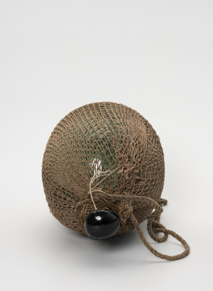 Pływak - ujęcie z góry; Wykonany z ciemnozielonego szkła pływak o kształcie przypominającym kulę. W górnej części pływak przechodzi w krótką, zakończoną spłaszczoną gałką szyjkę. Pływak w całości dwukrotnie owinięty siatką bawełnianą o małych, kwadratowych oczkach. Końce siatki ściśle przylegają do szyjki i są mocowane do niej za pomocą kilkukrotnie owiniętego wokół szyjki sznurka. Drugi, grubszy sznurek, służący jako mocowanie do sieci również owinięty jest wokół szyjki.