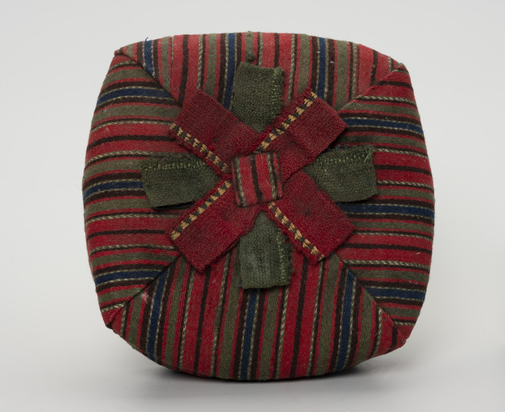 Czapka męska - ujęcie z góry; Czapka męska. Wykonana z tkaniny wełnianej, samodziałowej tzn. na krosnach tkackich splotem skośnym, wzór układa się w paski: czerwony, zielony, granatowy, brązowy, przetykany białym). Czapka ma fason biretu (zbliżonego do nakryć głowy duchownych m.in.  luterańskich). Uszyta jest z  czterech kawałków tkaniny usztywnionych od spodu i zszytych w ten sposób, że kształt zbliżony jest do kwadratu. Na czubku czerwono-zielona pasmanteryjna kokarda płasko ułożona w formę gwiazdy. Otok o zbliżonym do koła obwodzie z granatowej, wełnianej tkaniny.