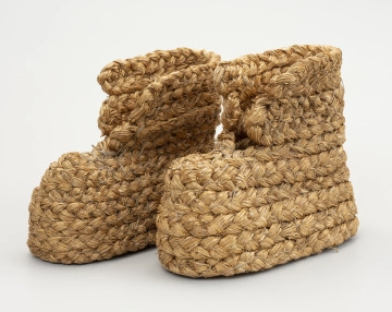 MNS/E/4496/a-b Buty słomiane - ujęcie z przodu; Buty słomiane wykonane techniką plecionkarską. Z uplecionego z powróseł słomy (prawdopodobnie żytniej) metodą spiralną warkocza wykonane buty o gładkiej podeszwie (bez obcasa), zabudowanej stopie oraz sięgającej połowy łydki prostej cholewce, z przodu rozchylonej. Poszczególne elementy spirali warkocza zszywane sznurkiem konopnym. Buty słomiane stosowane były do ocieplenia nóg w trakcie zimowych prac gospodarczo - rolniczych, jak np. w czasie młocki cepami.