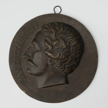 MNS/N/15910 Medalion z wizerunkiem Jana Kochanowskiego - ujęcie z przodu; Okrągły medalion z dolutowanym uchem i wizerunkiem głowy męskiej w lewo. Głowa poety z krótkimi falującymi włosami i długimi wąsami. Z lewej strony napis JAN KOCHANOWSKI.