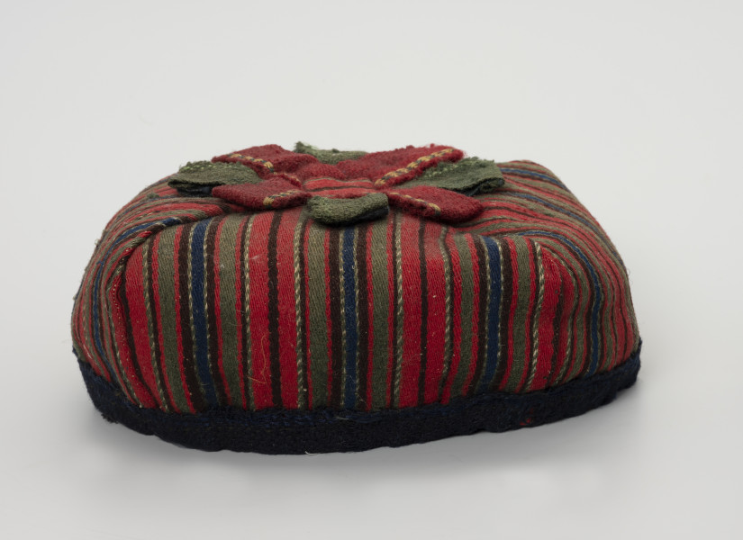 Czapka męska - ujęcie z boku; Czapka męska. Wykonana z tkaniny wełnianej, samodziałowej tzn. na krosnach tkackich splotem skośnym, wzór układa się w paski: czerwony, zielony, granatowy, brązowy, przetykany białym). Czapka ma fason biretu (zbliżonego do nakryć głowy duchownych m.in.  luterańskich). Uszyta jest z  czterech kawałków tkaniny usztywnionych od spodu i zszytych w ten sposób, że kształt zbliżony jest do kwadratu. Na czubku czerwono-zielona pasmanteryjna kokarda płasko ułożona w formę gwiazdy. Otok o zbliżonym do koła obwodzie z granatowej, wełnianej tkaniny.