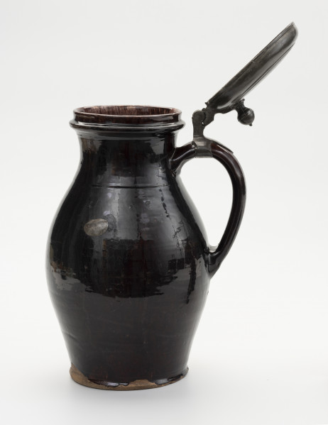 Dzban z pokrywą - ujęcie z przodu; Wysmukły dzban z otwartą pokrywą o lekko wyodrębnionej podstawie. Największa szerokość brzuśca przypada w połowie wysokości naczynia. Brzusiec łagodnym przewężeniem przechodzi w szeroką szyjkę; miejsce przejścia zaznaczone dookolną linią. Szyjka łagodnie przechodzi w wywinięty na zewnątrz wylew o zaoblonym rancie. Ucho szerokie, taśmowe, zaczepione pomiędzy dolną częścią szyjki, a  brzuścem poniżej najszerszej jego części. Na szczycie ucha obejma ze skierowaną pionowo do góry, ozdobnie profilowana kolumienką zaopatrzoną w zawias, służący do przymocowania pokrywy, szczelnie zamykającej wylew. Pokrywka z bolcem w postaci kolumienki zakończonej kopułką z małą kulką na szczycie, tworzące paluch do jej podnoszenia. Dzban  w całości pokryty szkliwem, w środku cieńszą warstwą a na zewnątrz grubszą. Naczynie z wyraźnymi śladami naprawy: dziura w brzuścu zalepiona cynową, lutowana łatą, widoczne ślady po odrutowaniu, utrzymującym w całości pękniętą konstrukcję.