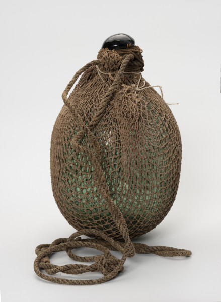 Pływak - ujęcie z przodu; Wykonany z ciemnozielonego szkła pływak o kształcie przypominającym kulę. W górnej części pływak przechodzi w krótką, zakończoną spłaszczoną gałką szyjkę. Pływak w całości dwukrotnie owinięty siatką bawełnianą o małych, kwadratowych oczkach. Końce siatki ściśle przylegają do szyjki i są mocowane do niej za pomocą kilkukrotnie owiniętego wokół szyjki sznurka. Drugi, grubszy sznurek, służący jako mocowanie do sieci również owinięty jest wokół szyjki.